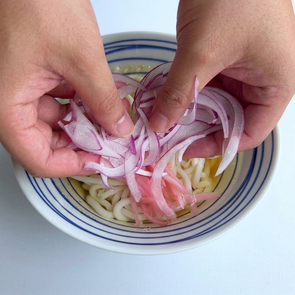 Two hands dropping a pile of thinly sliced purple-red onions onto the center of a bowl of udon noodles.