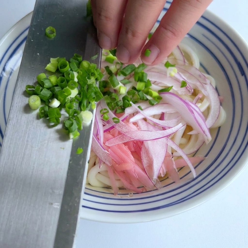 Finely chopped green onions being scraped off the side of a metal knife onto a bowl of udon noodles with sliced red onions.