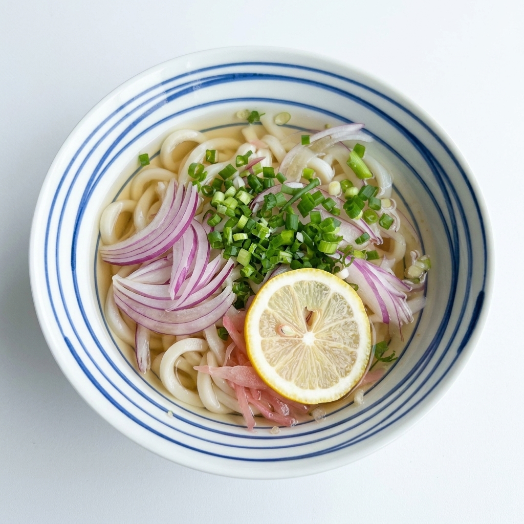 An overhead view of a bowl of udon noodles in clear broth, topped with red onions and scallions, with a single yellow lemon slice placed in the middle.