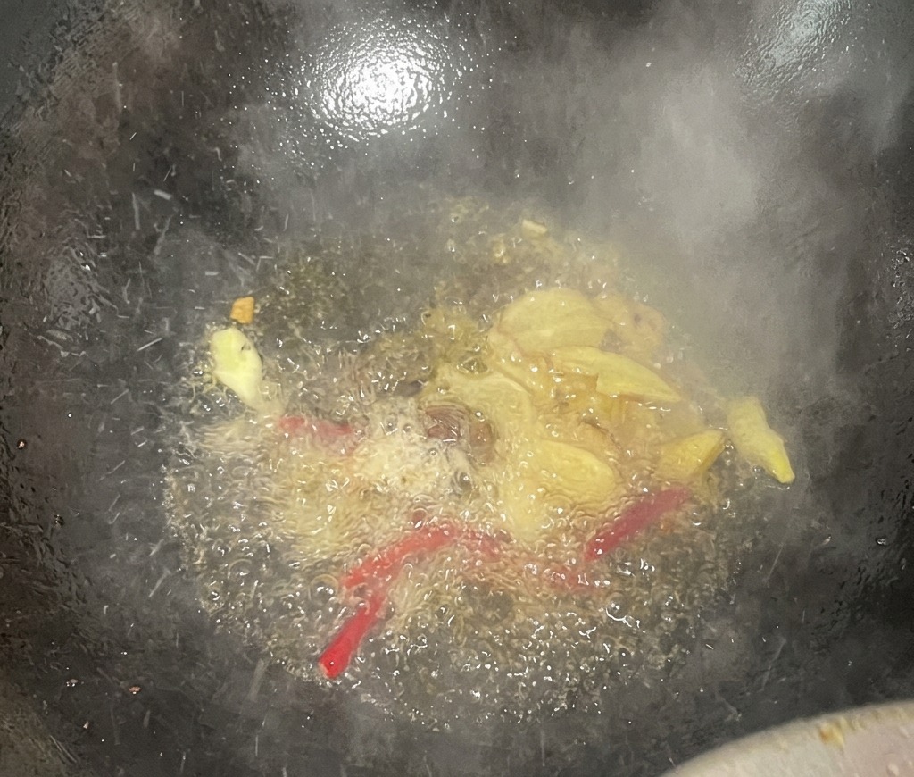 Ginger slices and red dried chilies bubbling in hot oil inside a dark wok, releasing steam and aromatics.