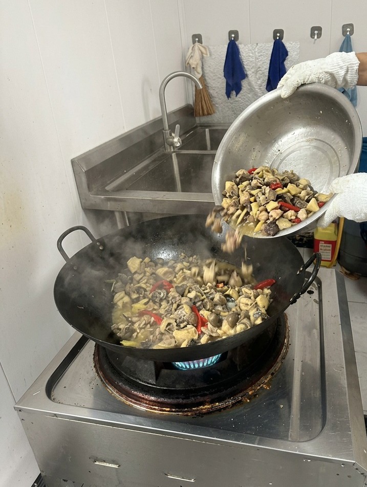 A person pouring clear boiling water from a vessel into a steaming wok containing seared chicken and spices.