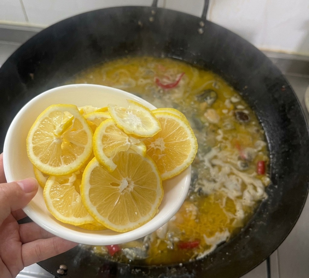 A hand holding a white bowl of thin lemon slices over a bubbling wok filled with golden chicken broth.