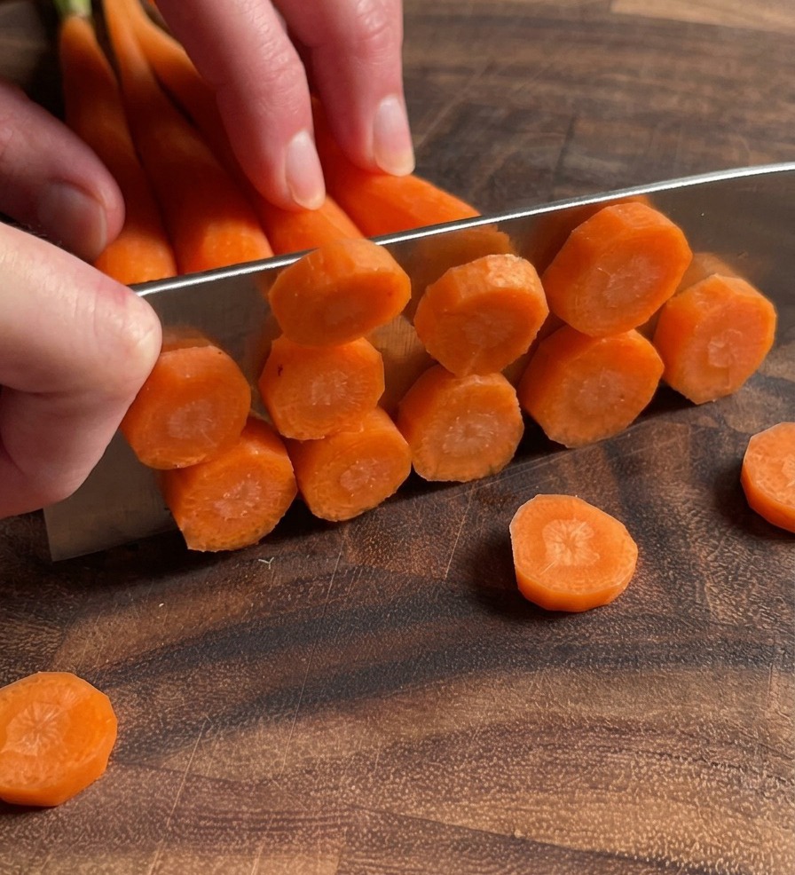 A close up shot of hands using a knife to slice multiple carrots into thin rounds on a dark wooden cutting board.