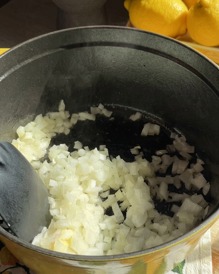 Diced white onions being stirred in a dark cooking pot as they begin to soften in butter.