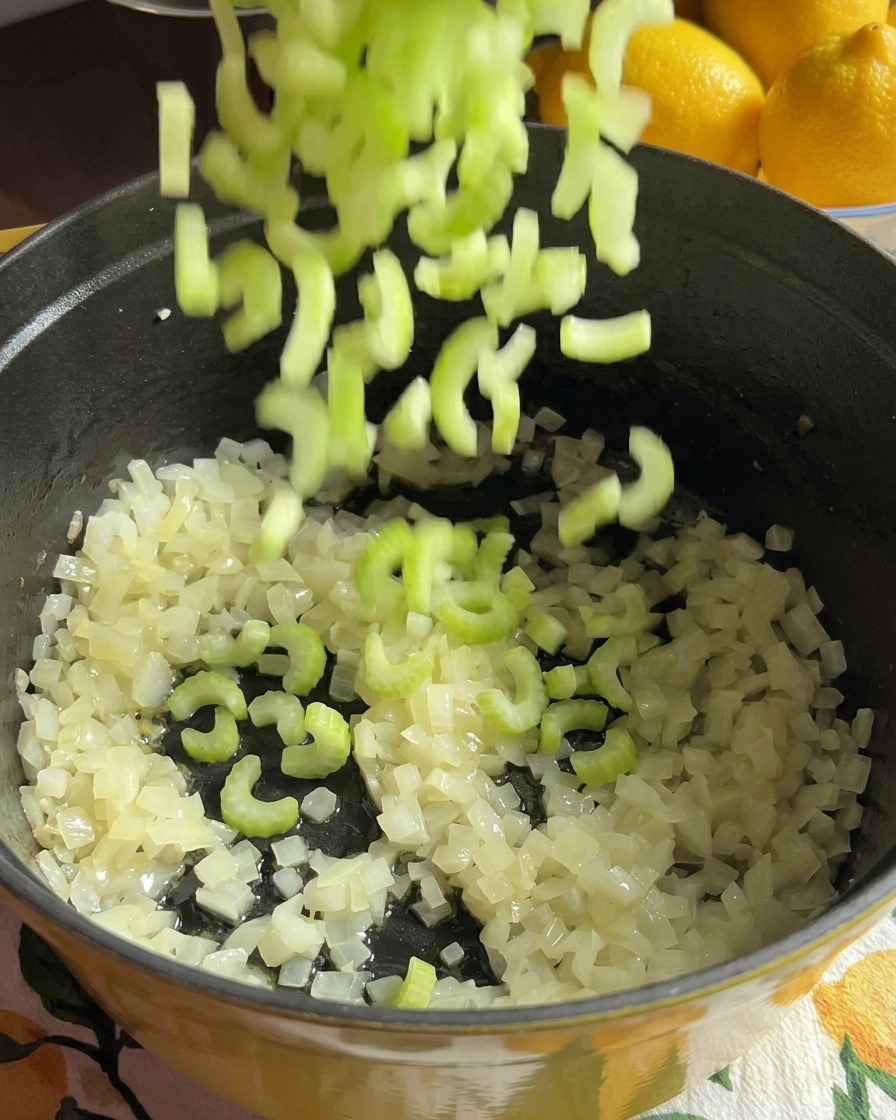 Chopped green celery pieces falling into a cooking pot containing onions and sliced carrot rounds.