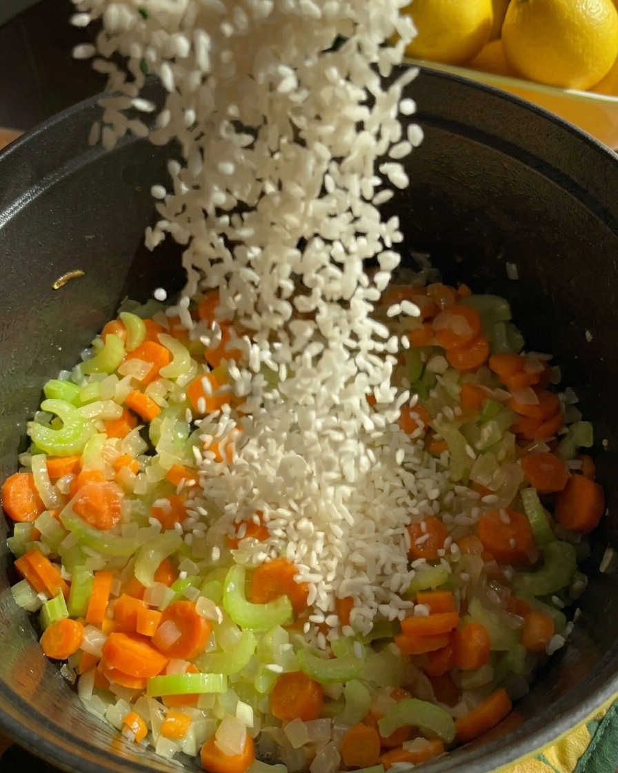 A stream of white jasmine rice being poured into a dark pot filled with sautéed diced carrots, celery, and onions.