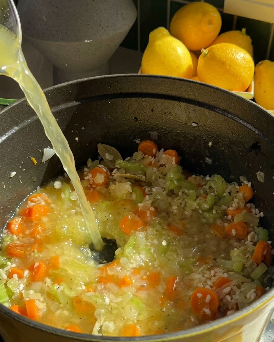 A stream of clear chicken broth being poured into a pot containing rice, carrots, and celery.