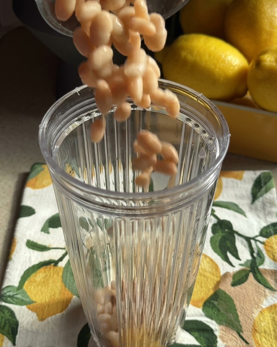 Cooked white beans falling into a clear blender container sitting on a lemon-patterned kitchen towel.