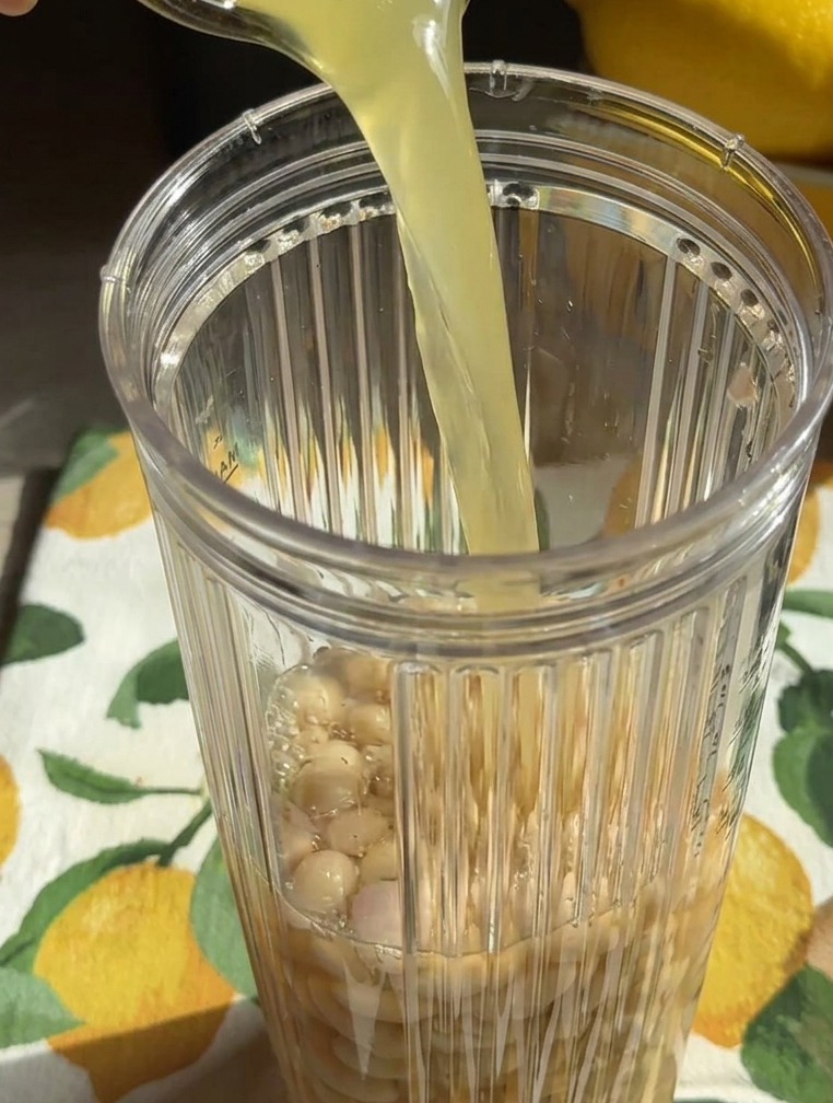 A yellowish liquid being poured from a glass into a blender cup filled with whole white beans.