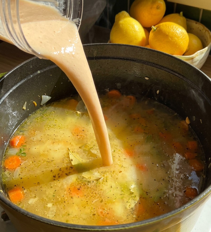 A stream of thick, creamy beige bean puree being poured from a blender cup into a pot of simmering soup with sliced carrots.