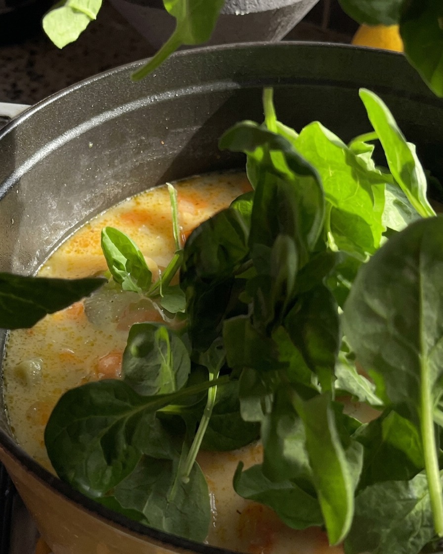 Handfuls of fresh green baby spinach leaves being dropped into a pot of creamy soup.