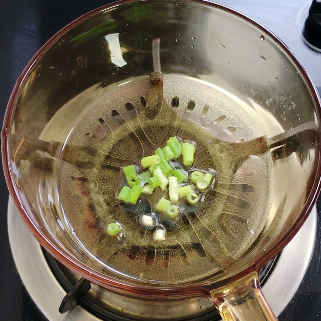 Finely chopped green scallions simmering in a pool of clear oil at the bottom of a transparent glass cooking pot.