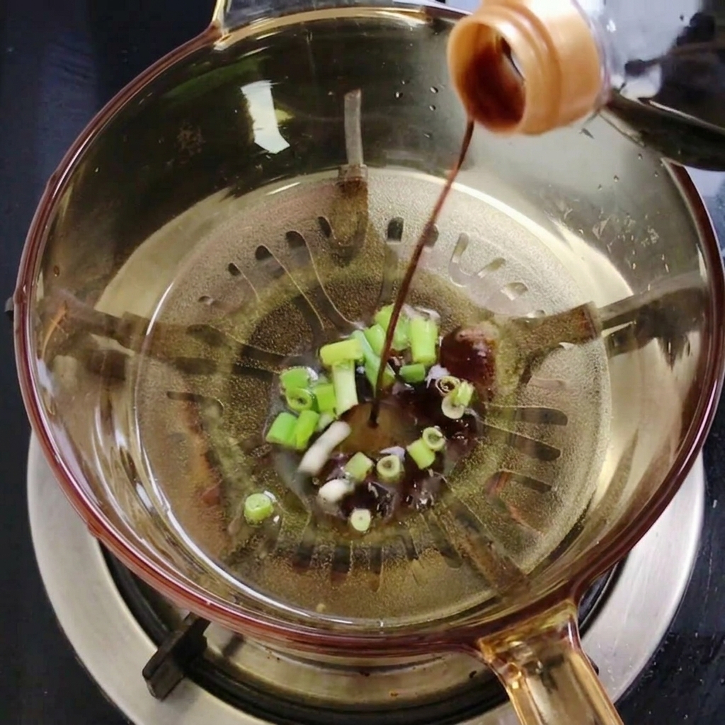 A stream of dark soy sauce being poured from a bottle into a glass pot containing sautéed green onions and oil.