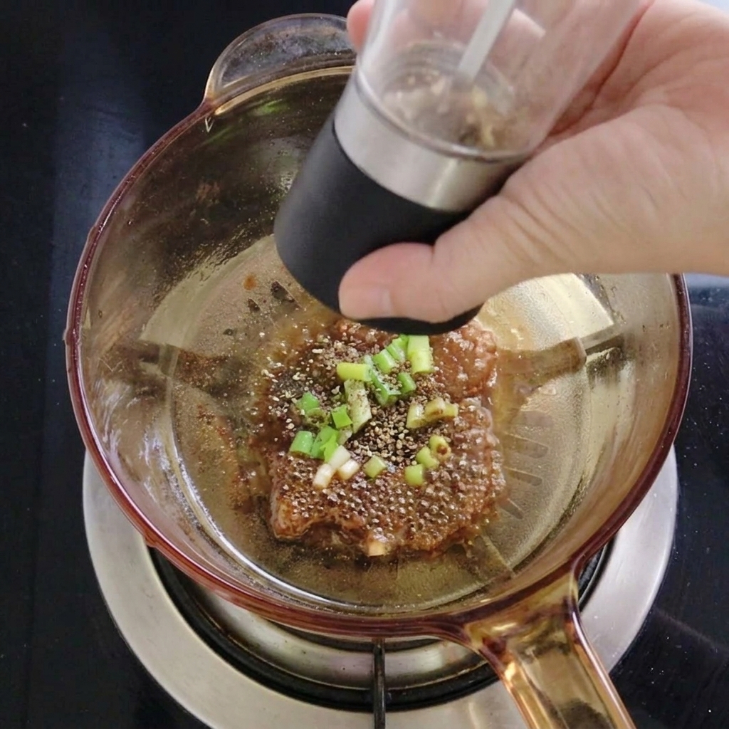 A person using a black manual grinder to sprinkle ground peppercorn powder over the oil and scallion mixture in a glass pot.
