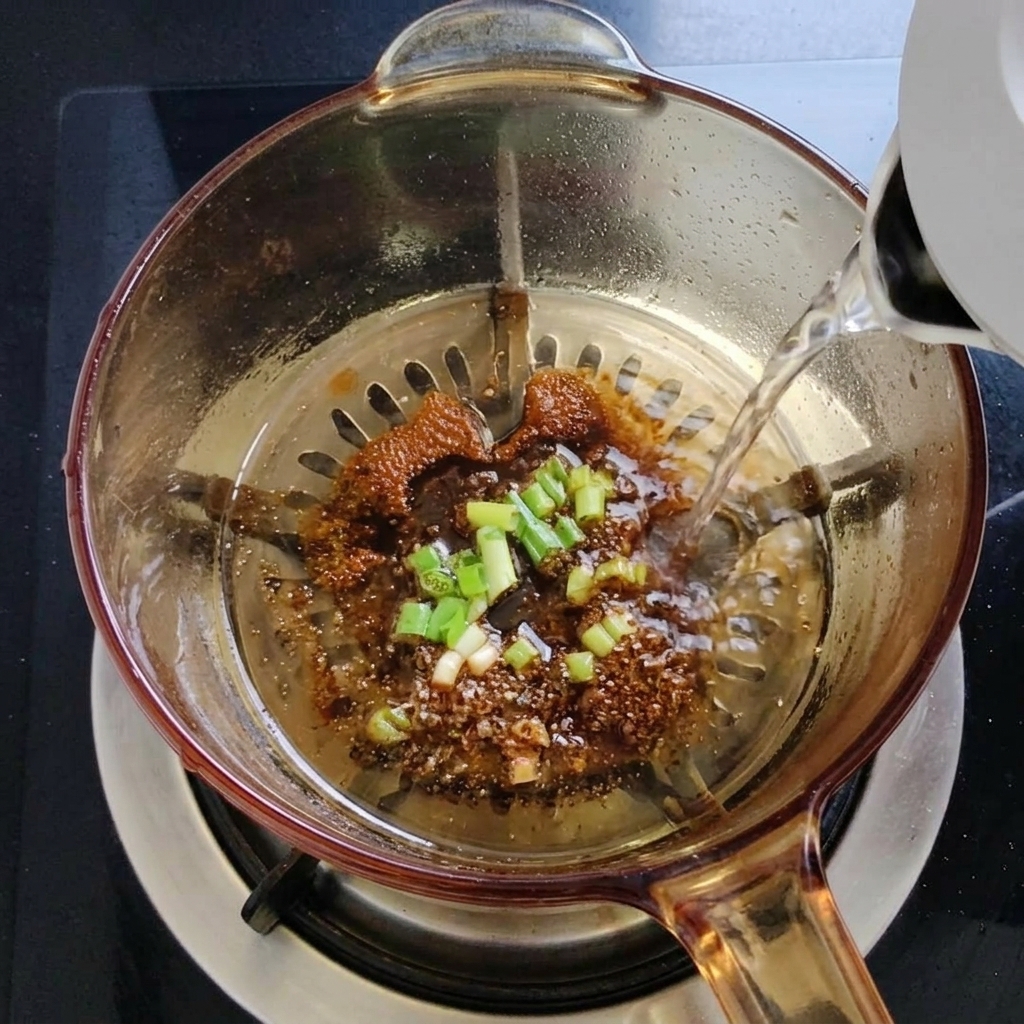 Hot water being poured from a white electric kettle into a glass pot, creating steam as it hits the seasoned oil base.