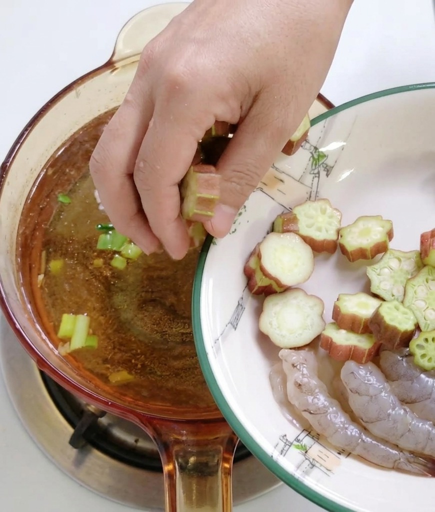 A persons hand adding sliced green okra rounds from a white plate into a bubbling brown soup base in a glass pot.