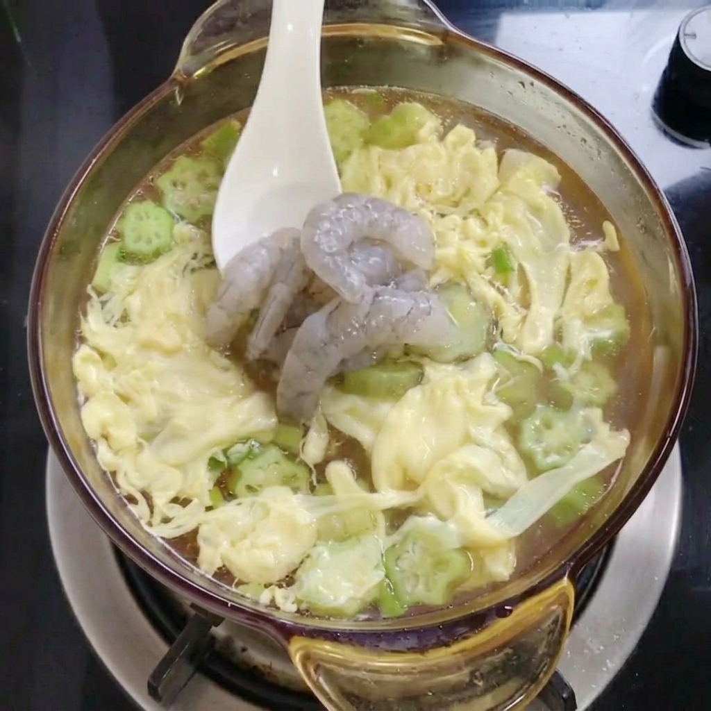 A white spoon placing several raw grey shrimp into a glass pot filled with yellow egg ribbons and green okra slices in broth.