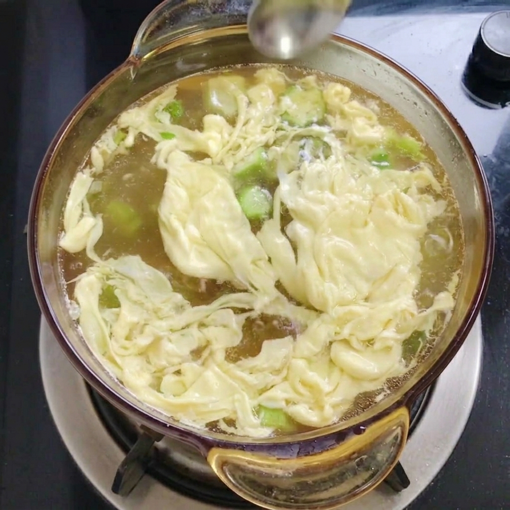 A metal spoon sprinkling fine white salt into a pot of boiling soup containing egg ribbons and okra.