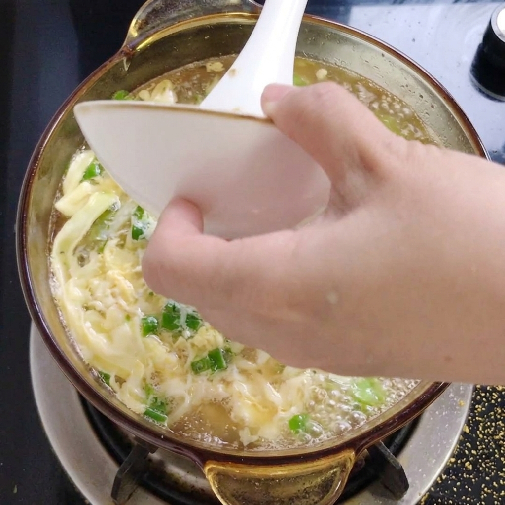 Chopped green herbs being poured from a small white bowl into a steaming pot of okra and egg soup.
