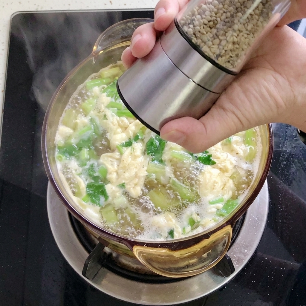 A hand using a stainless steel pepper grinder to add white pepper to a pot of soup filled with greens and egg ribbons.