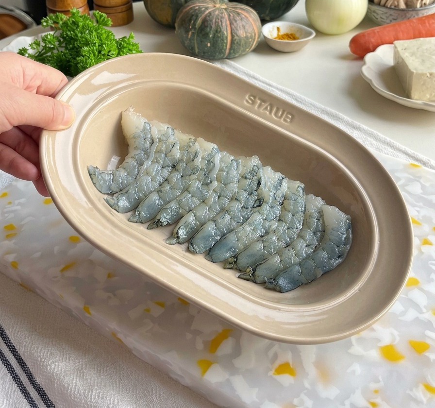 Freshly de-veined raw shrimp being seasoned in a beige oval ceramic dish with salt and pepper grinders visible in the background.