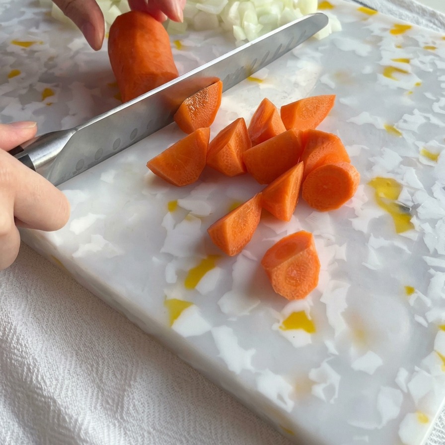 A kitchen knife slicing a bright orange carrot into chunks on a white and yellow patterned cutting board next to a pile of diced onions.