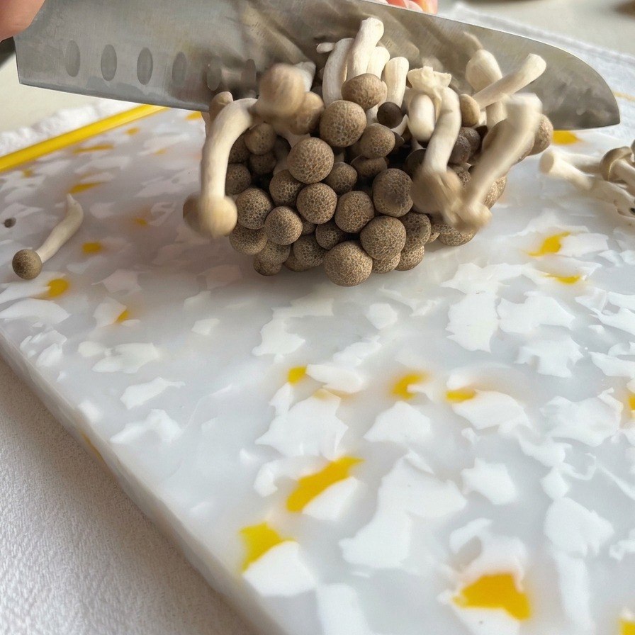 A chefs knife slicing through the base of a cluster of brown shimeji mushrooms on a patterned cutting board.