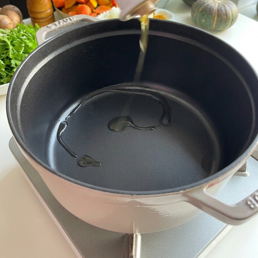 Clear cooking oil being poured from a white ceramic bottle into a preheated black Staub cast iron pot.
