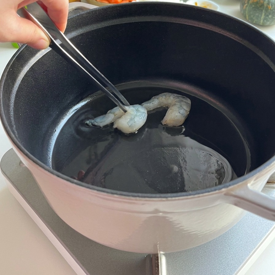 Raw shrimp being placed into a black pot with metal tweezers for searing in hot oil.