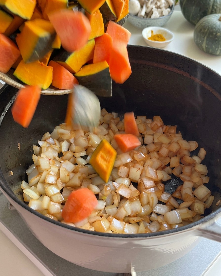 Fresh pumpkin chunks and cut carrots being added to sautéed onions in a pot.