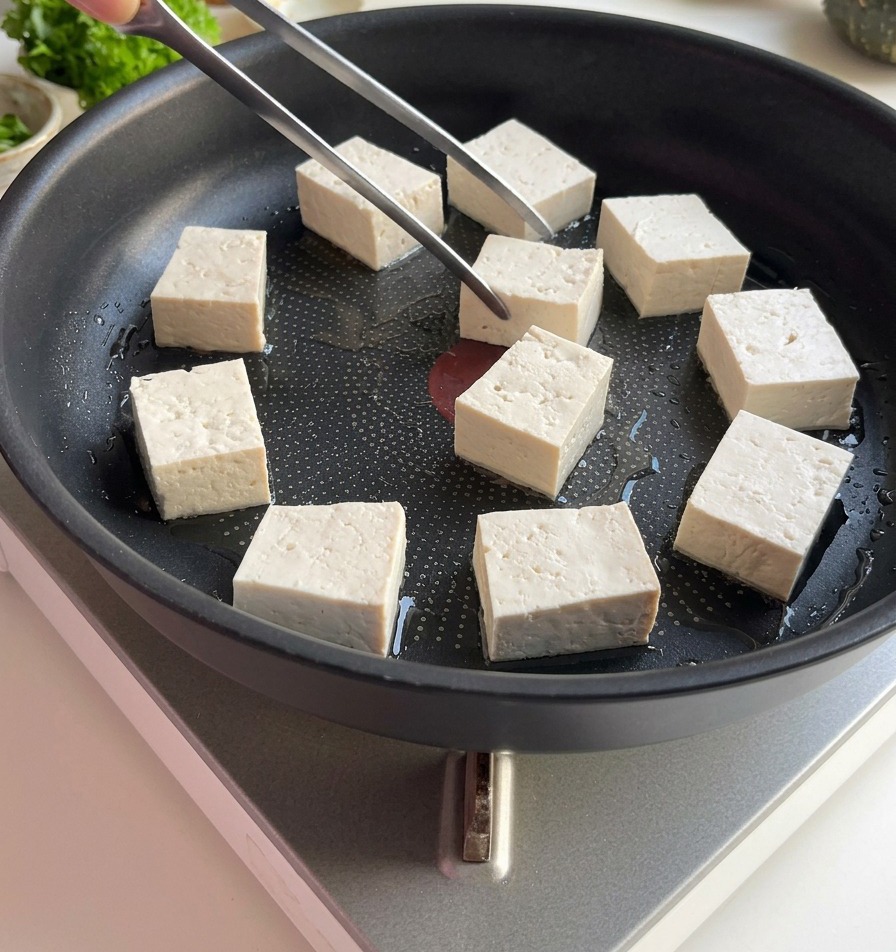 Tweezers placing white, square tofu blocks into a flat frying pan with a light coating of oil.