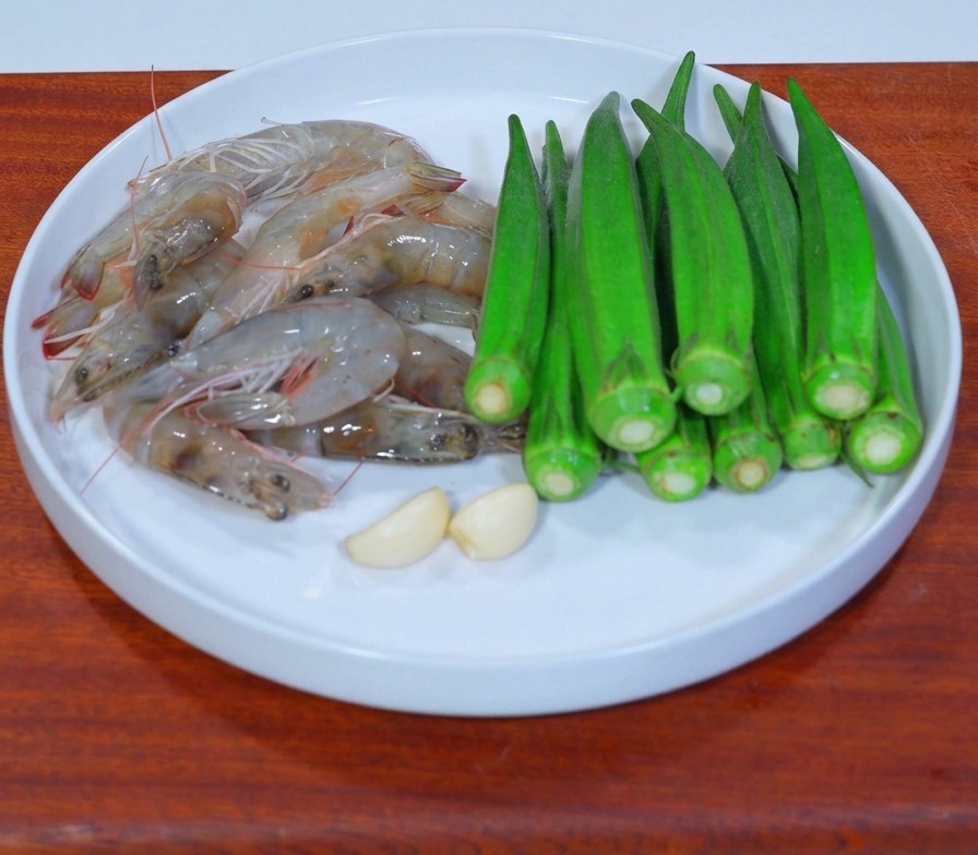 A white plate displaying fresh raw prawns, green okra pods, and two garlic cloves on a wooden surface.