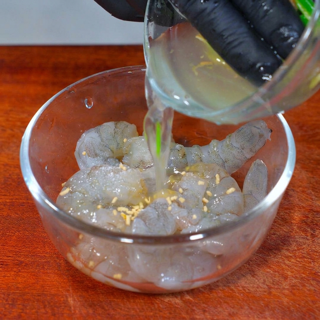 Aromatic scallion and ginger water being poured into a glass bowl of seasoned raw shrimp.