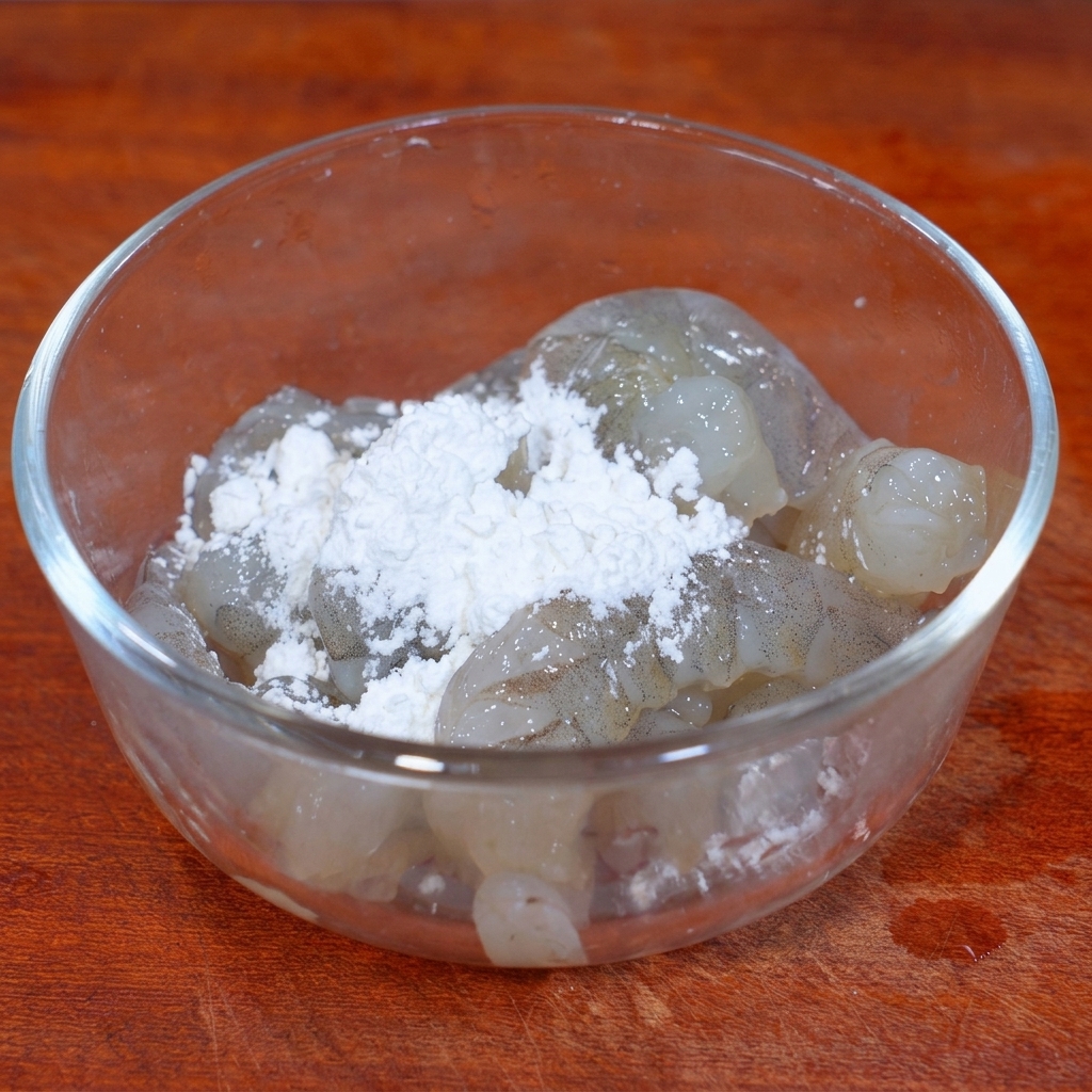 A pile of white starch powder sitting on top of marinated raw shrimp meat in a clear bowl.