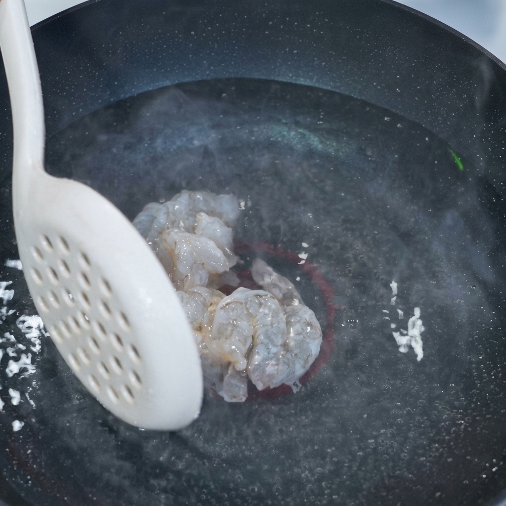 A white slotted spoon holding several pieces of translucent, marinated shrimp meat over a pot of steaming water.