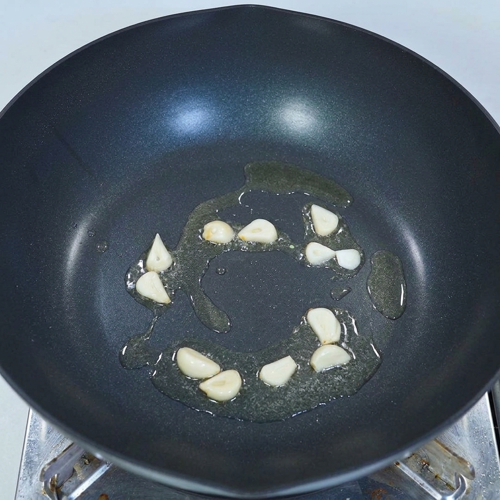 Thinly sliced garlic cloves sizzling in a shallow pool of oil at the bottom of a dark wok.