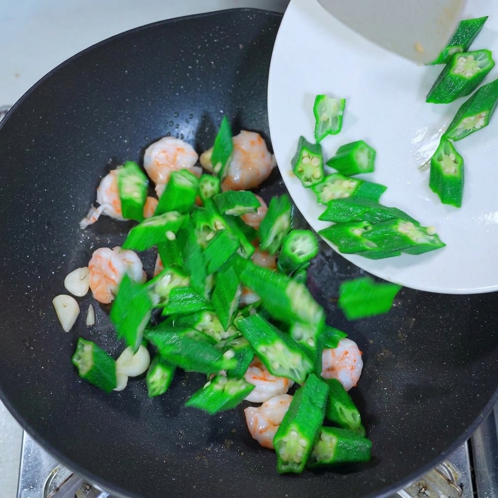 Bright green okra slices being poured from a white plate into a wok containing cooked shrimp and garlic slices.