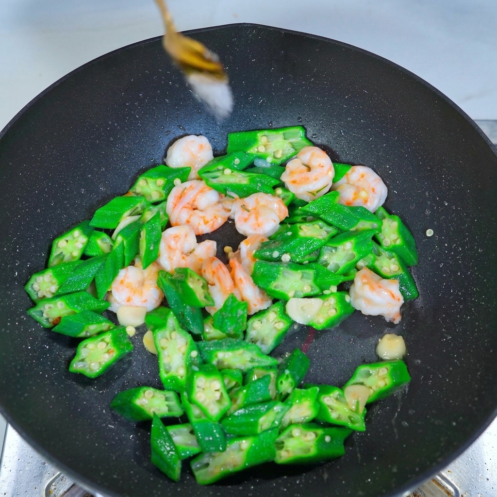 A metal spoon sprinkling white seasoning over a mixture of bright green okra slices and pink shrimp in a wok.