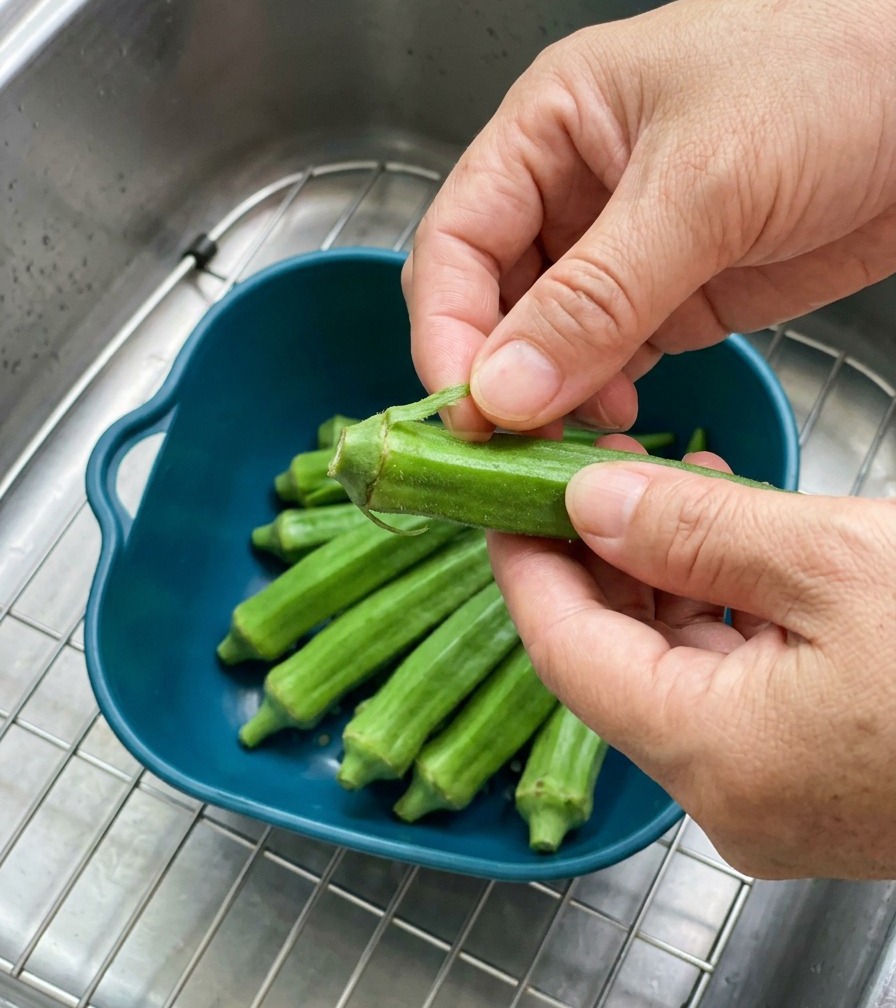 A persons hands holding a vibrant green okra pod, pointing out the intact sepals at the stem end over a bowl of fresh okra.