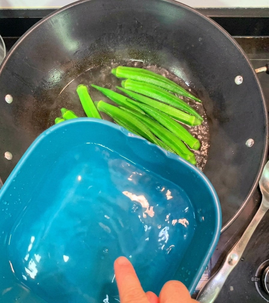 Bright green blanched okra pods being moved from a steaming wok into a nearby basin of cold water.
