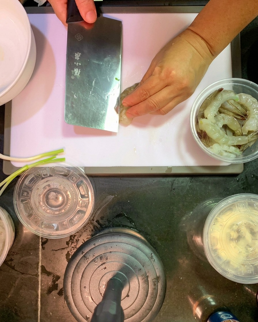 A kitchen knife making a shallow incision along the back of a raw, peeled shrimp on a white plastic cutting board.