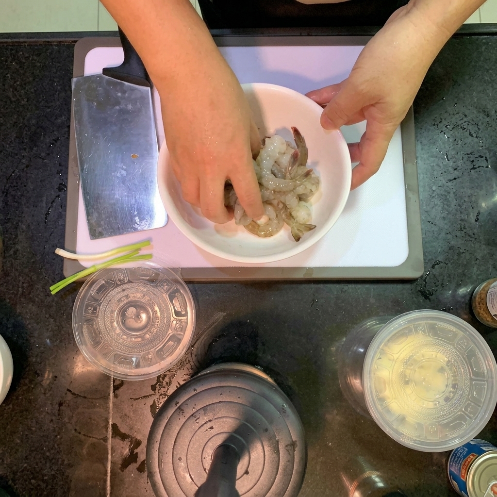 Hands mixing raw, translucent shrimp with salt and black pepper in a white ceramic bowl.