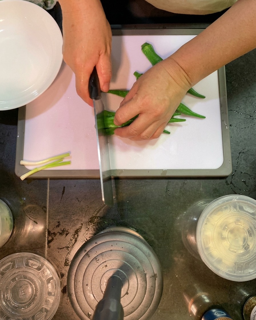 A knife slicing through blanched okra pods at a diagonal angle on a white cutting board.