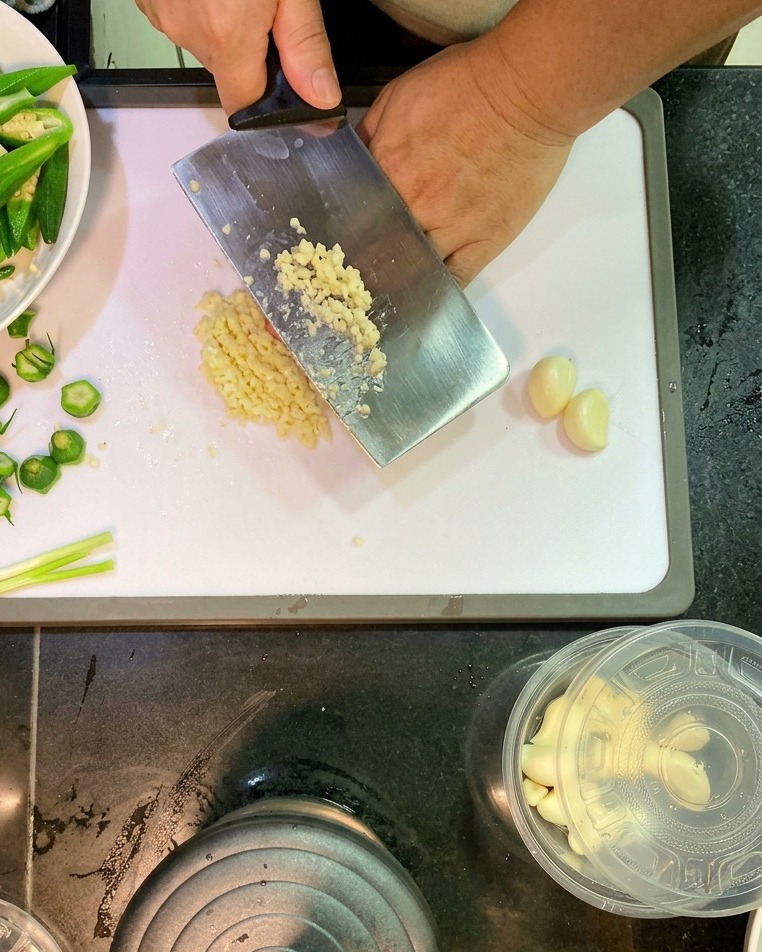 A close-up shot of a large metal cleaver mincing a pile of white garlic cloves into small bits on a white cutting board.