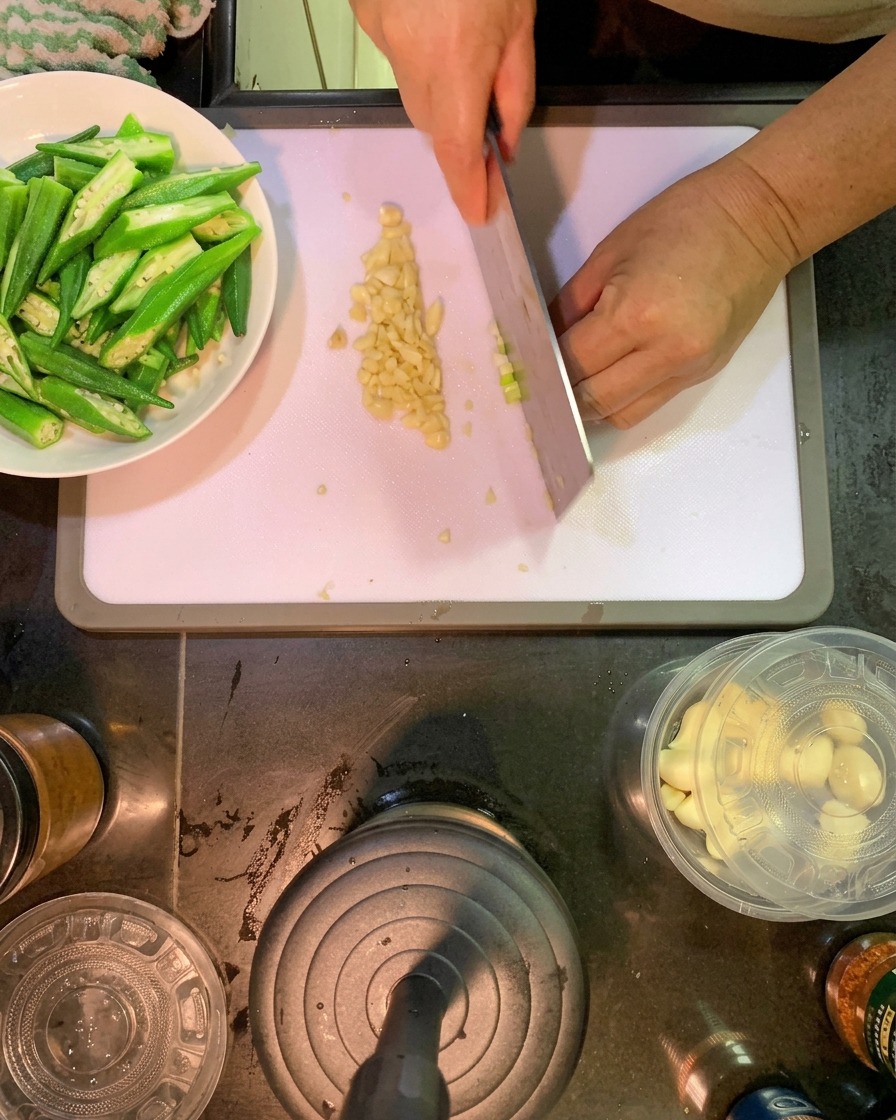 A person using a large kitchen knife to slice the white parts of green onions and mince garlic on a white cutting board, with sliced okra in a side dish.