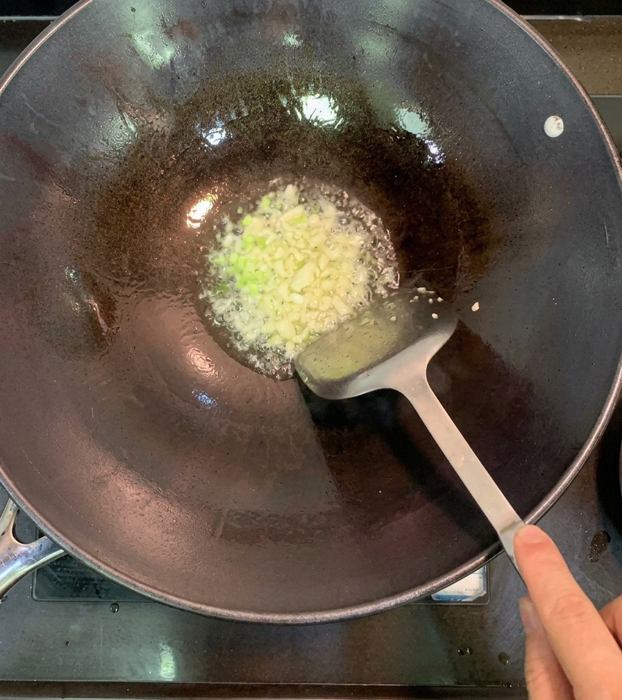 A top-down view of minced garlic and chopped green onions sizzling in hot oil at the bottom of a dark metal wok.