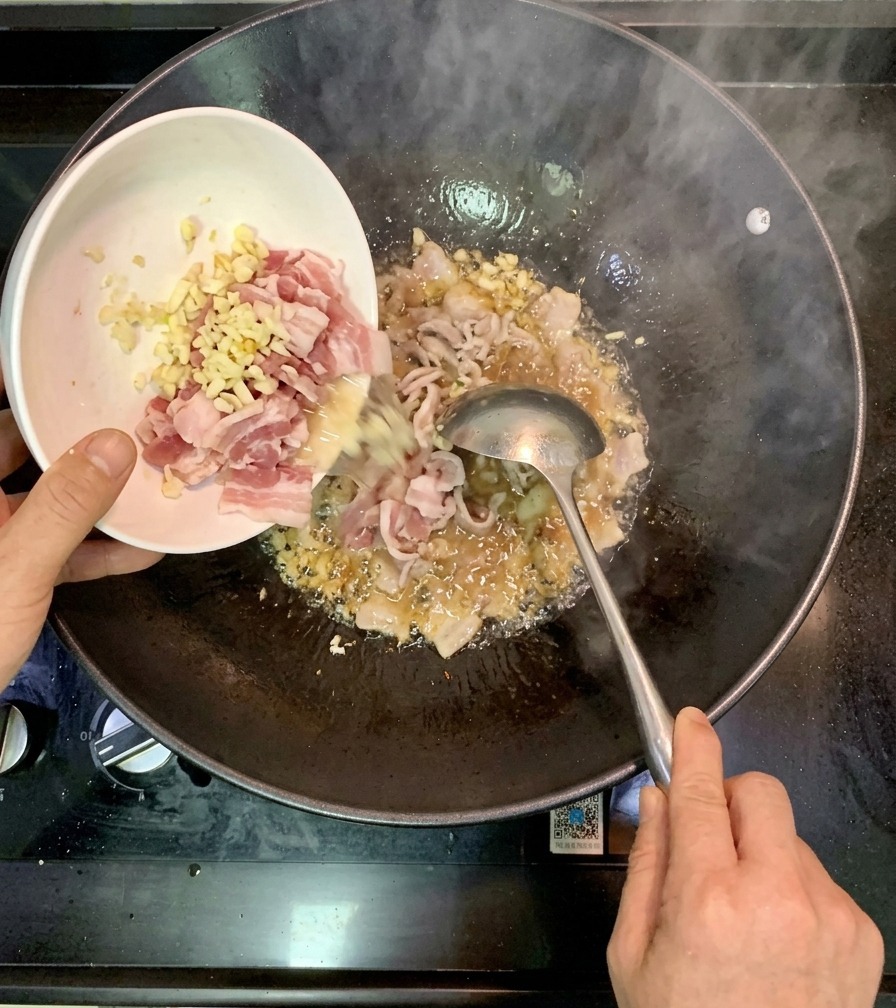 A person pouring pieces of marinated raw shrimp from a small white bowl into a hot wok containing sautéed garlic.