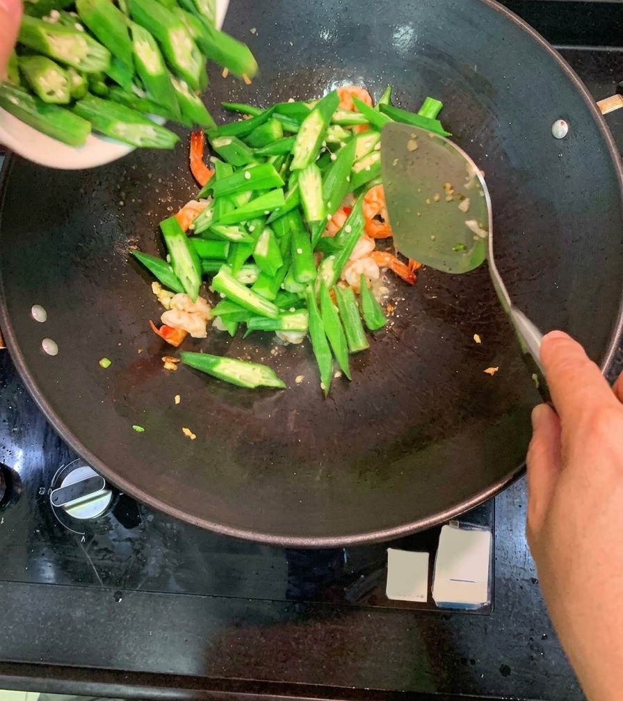 A bowl of sliced green okra being poured into a wok that already contains cooked pink shrimp and aromatics.