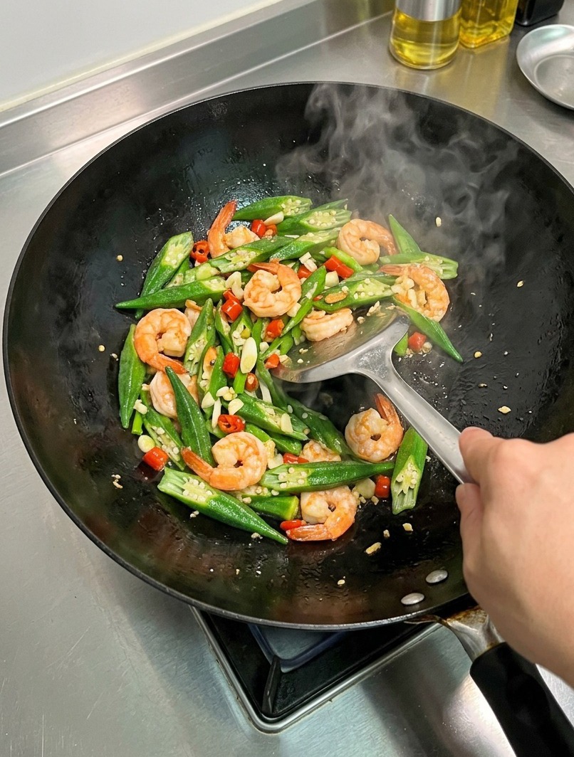 A close-up of a wok filled with stir-fried okra, shrimp, and red chili peppers being tossed with a metal spatula.