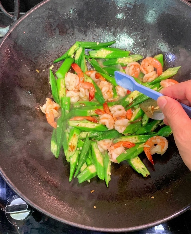 A person uses a small blue plastic spoon to add a pinch of salt to a stir-fry of shrimp and sliced okra in a black wok.